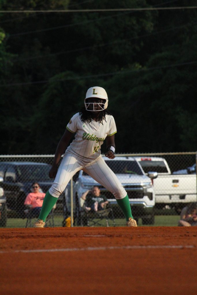 A softball player in uniform stands ready on the field, with spectators and vehicles visible in the background.