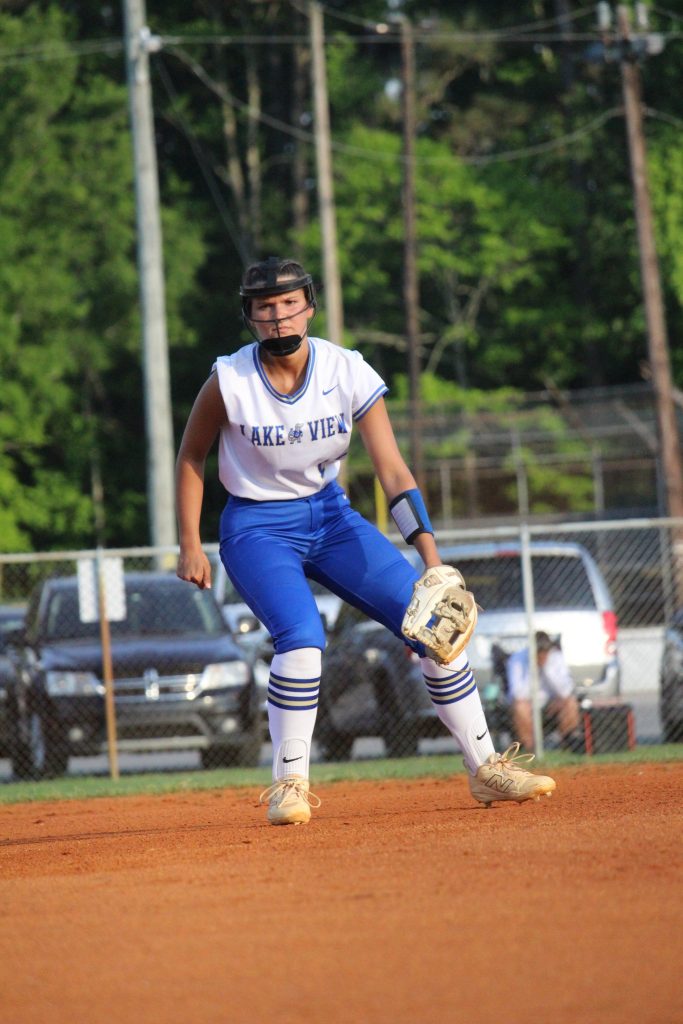 Softball player in blue pants and white jersey stands ready on the infield, wearing a face guard and glove, with cars and trees in the background.