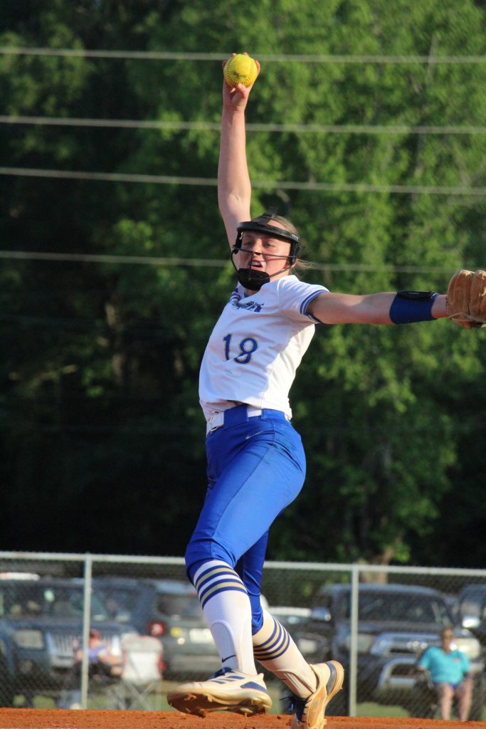 A softball player in blue and white uniform pitches the ball during a game, with trees and a fence in the background.