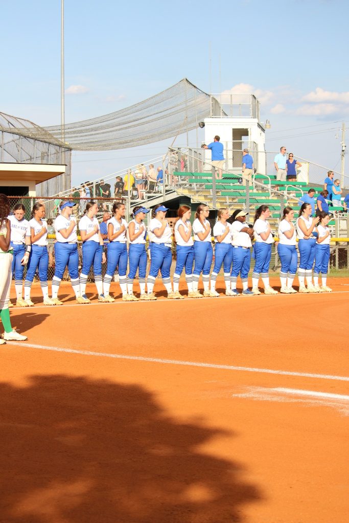 A softball team in blue and white uniforms stands in a line on the field, holding their hands over their hearts during the national anthem.