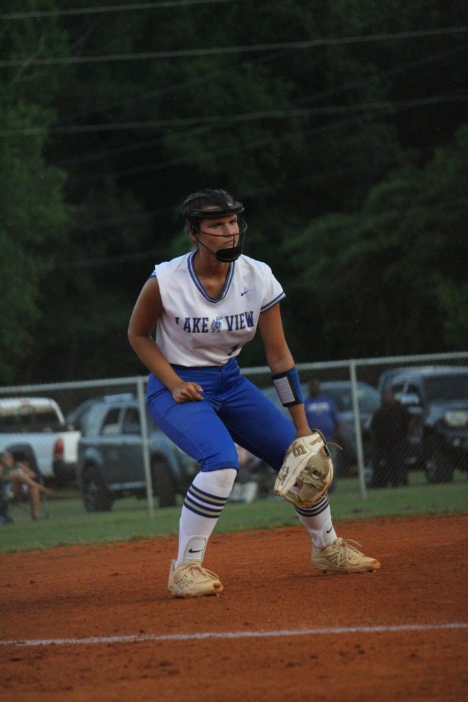 A softball player in uniform is ready in a fielding position on a dirt infield, with trees and parked cars in the background.