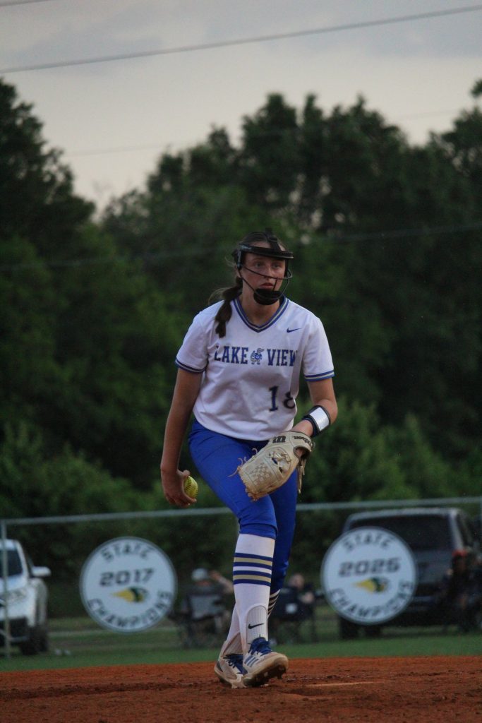 A softball player wearing a helmet and uniform is preparing to pitch on a field, with trees and signs visible in the background.