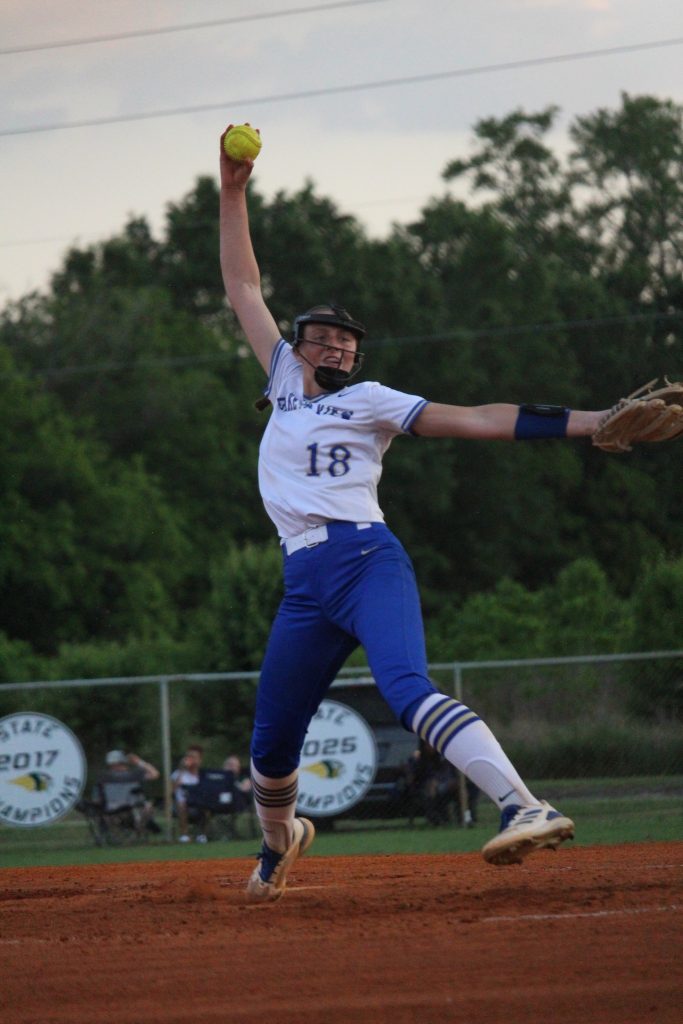 A softball player in mid-pitch wearing a white and blue uniform, numbered 18, on a field with trees and signs in the background.