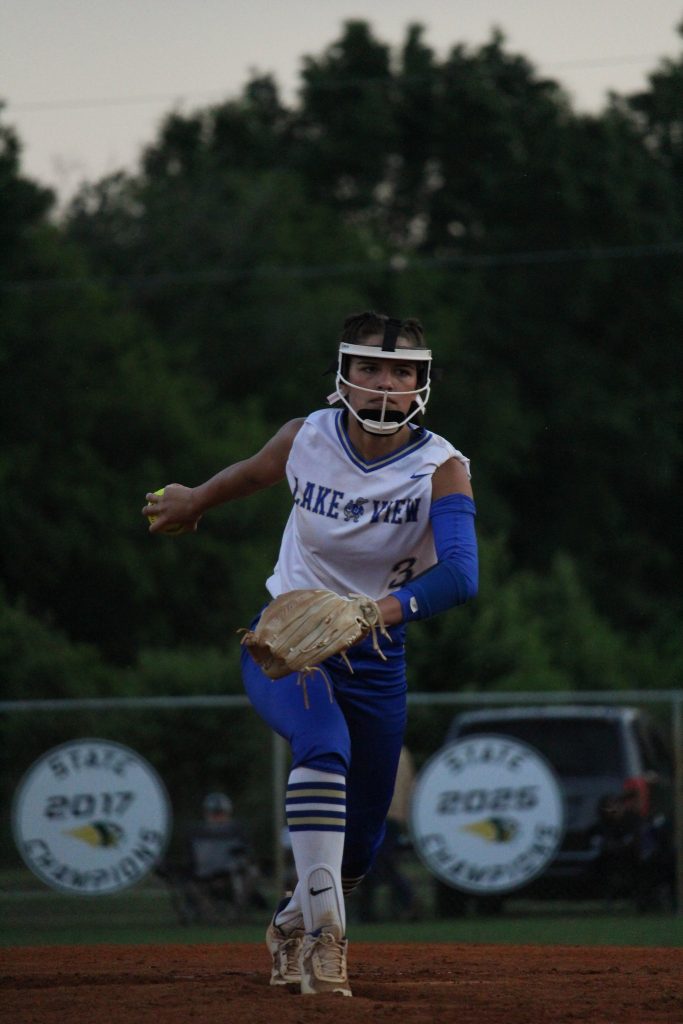 A softball player in a white and blue uniform runs on the field, wearing a helmet and glove, with trees and banners visible in the background.