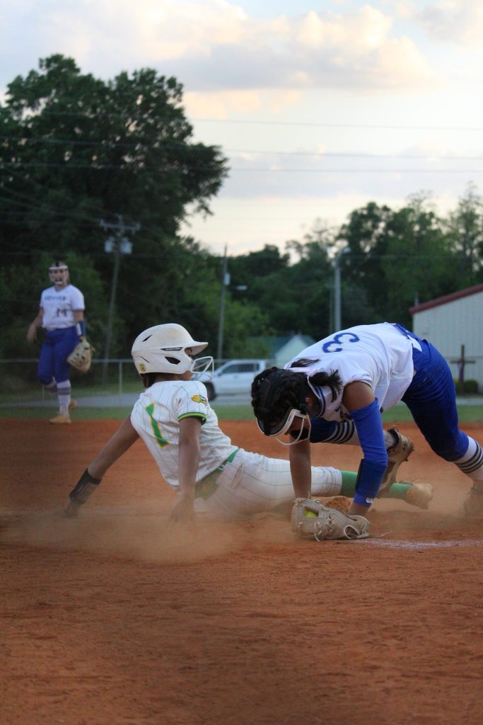 A baseball player slides into a base as an opposing player attempts to tag them, with dust rising from the ground and another player in the background.