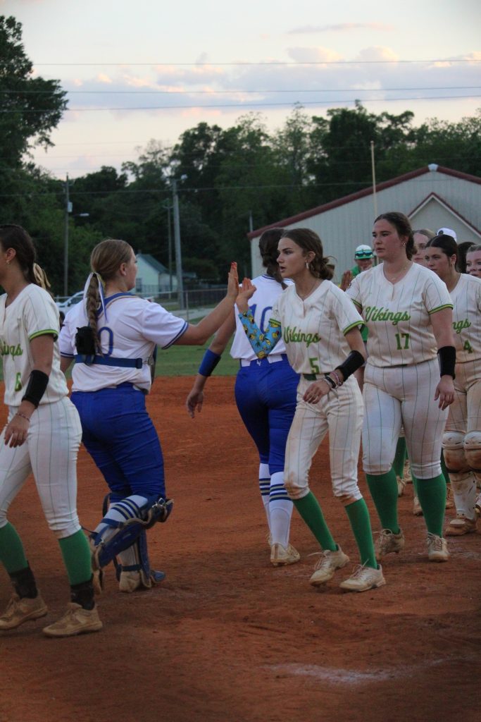 Softball players in uniforms high-fiving each other on a dirt field after a game, with trees and buildings in the background.