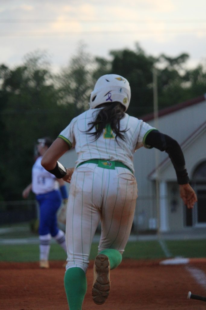 A softball player in a white uniform runs on a field, with a building and another player in the background.