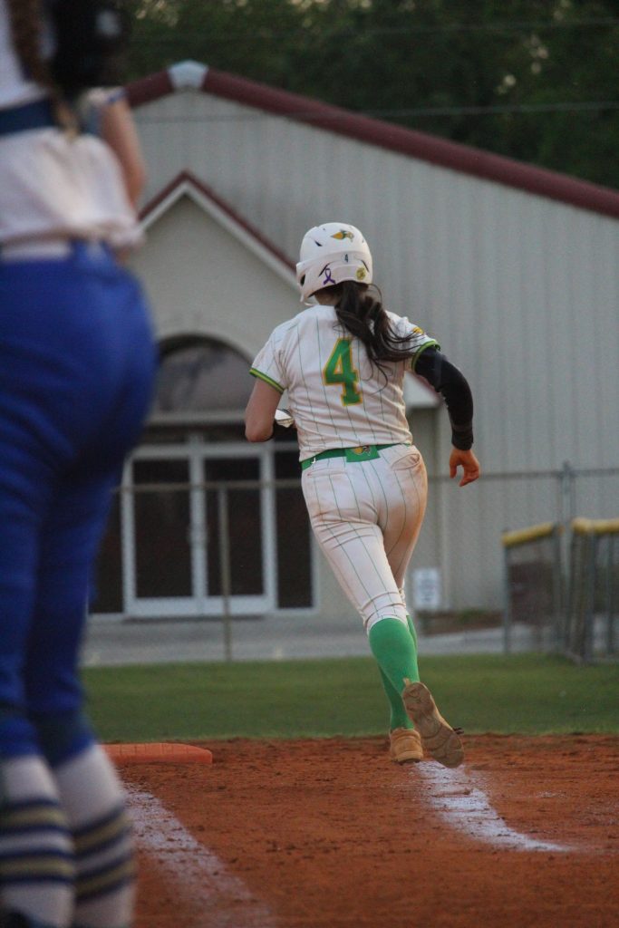 A softball player in a white and green uniform with number 4 runs during a game, while another player in blue stands nearby.