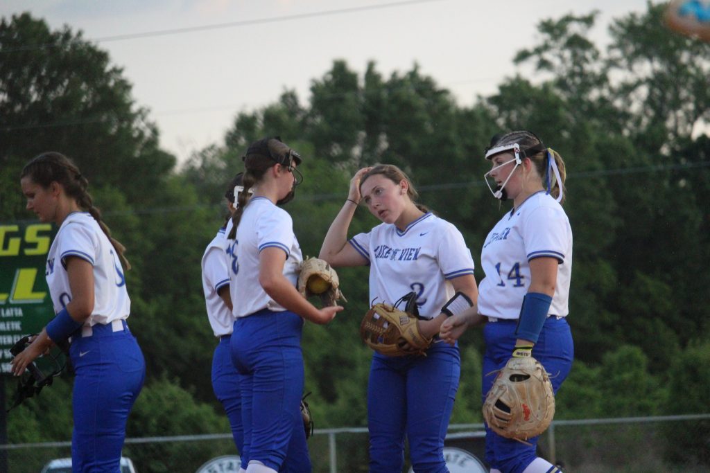 Four female softball players in white and blue uniforms stand together on a field, talking and holding gloves, with trees in the background.
