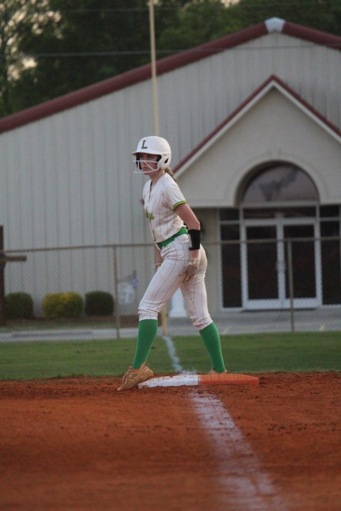 A softball player in uniform and helmet stands on a base on a dirt field, with a building and greenery in the background.