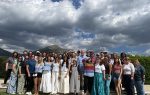 A large group of people standing outdoors on a sunny day with mountains and dramatic clouds in the background.