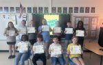 A group of students in a classroom proudly hold certificates, posing for a photo together. An American flag, posters, and a "Student Awards" screen are visible behind them.