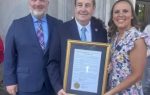 Three people stand outside, smiling at the camera. The man in the center holds a large framed certificate.