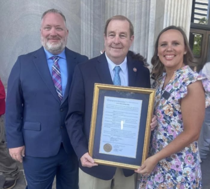 Three people stand outside, smiling at the camera. The man in the center holds a large framed certificate.
