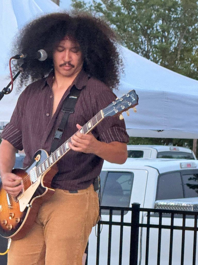 A musician with voluminous curly hair playing an electric guitar outdoors, standing near a microphone, with white tents and vehicles in the background.
