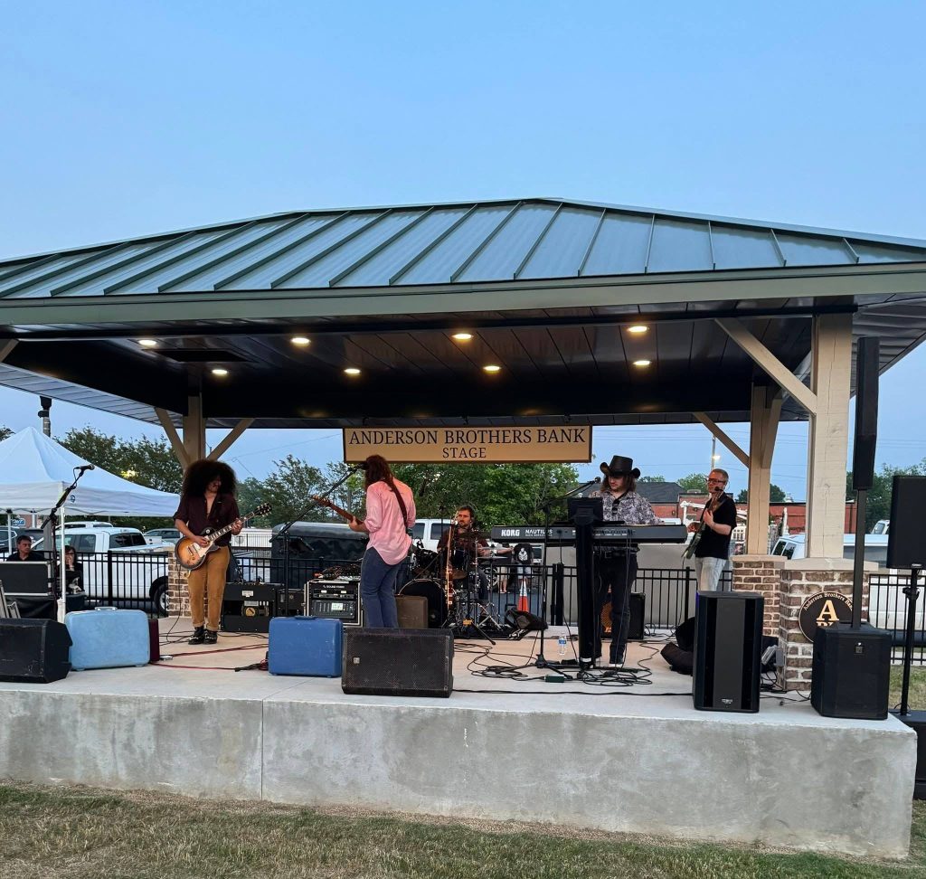 A band performs on an outdoor stage under a metal roof, with musicians playing guitar, keyboard, bass, and drums in front of an audience.