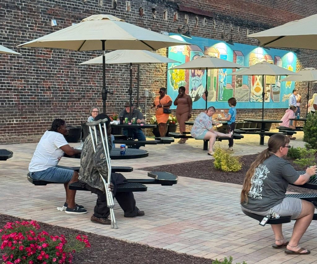 People sitting at outdoor tables under umbrellas near a brick wall with a colorful mural, enjoying a casual gathering on a paved patio with flower beds.