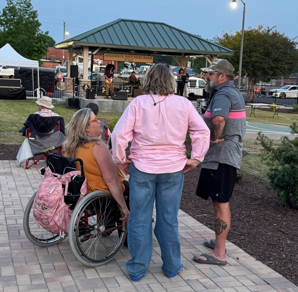 Three people converse near a wheelchair at an outdoor concert, with a band performing on a stage under a pavilion labeled "Anderson Brothers Bank Stage."