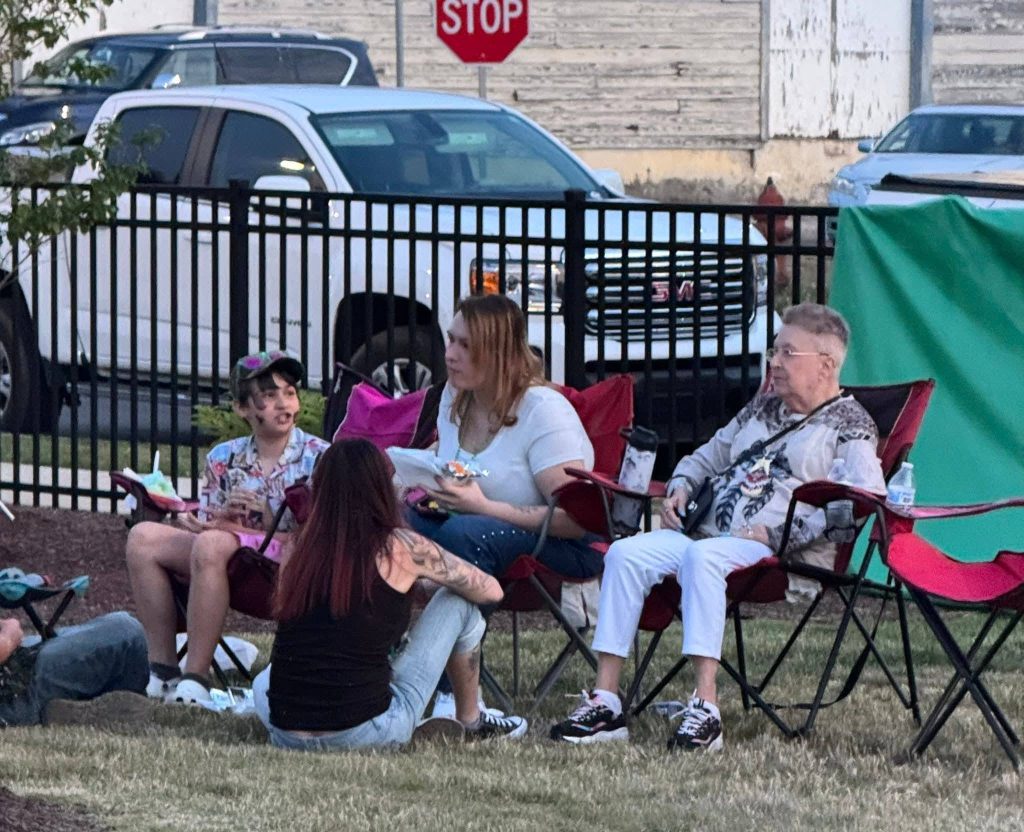 A group of people sitting on chairs and grass outdoors, eating and talking near a black fence with parked cars and a stop sign in the background.