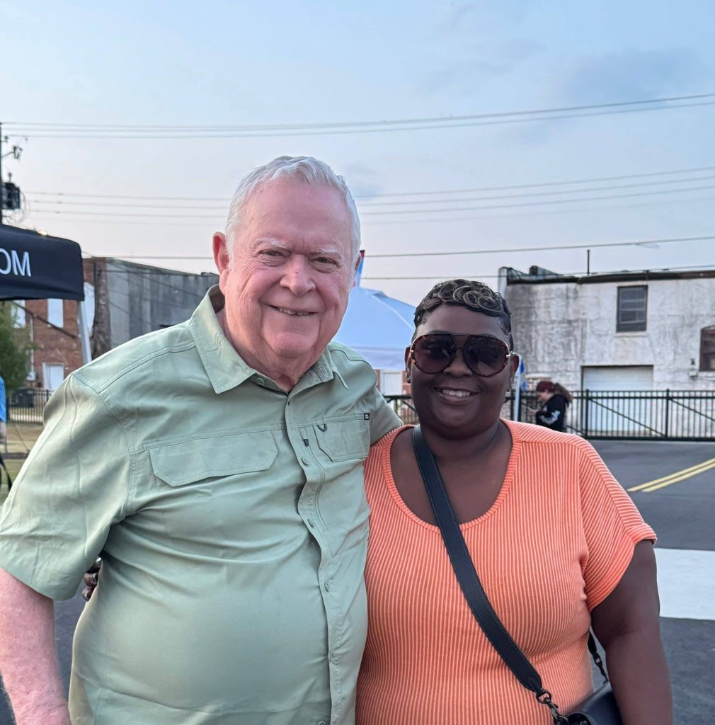 older man and a woman smiling and posing together outdoors at an event, with buildings and tents in the background.