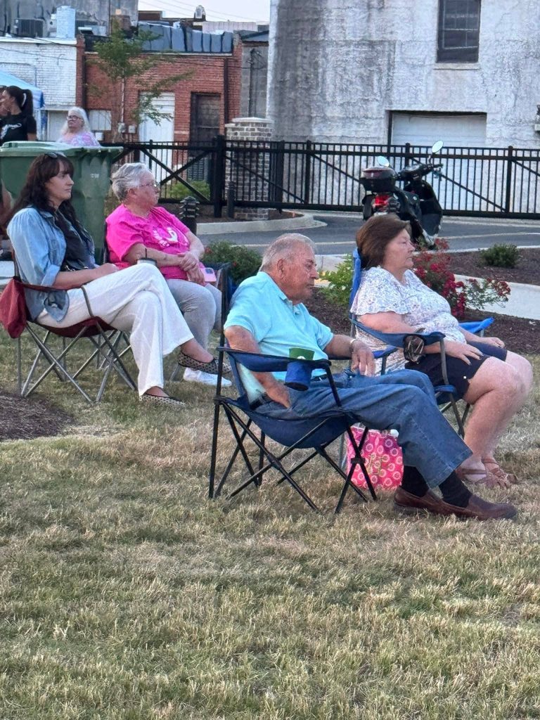 Four adults sit in portable chairs on grass, watching an outdoor event with buildings and a motorcycle visible in the background.