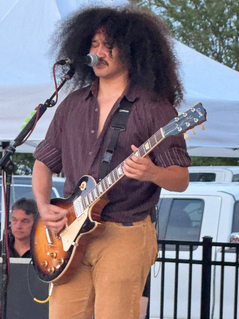 Man with curly hair playing an electric guitar and singing into a microphone on an outdoor stage, with tents and a person in the background.