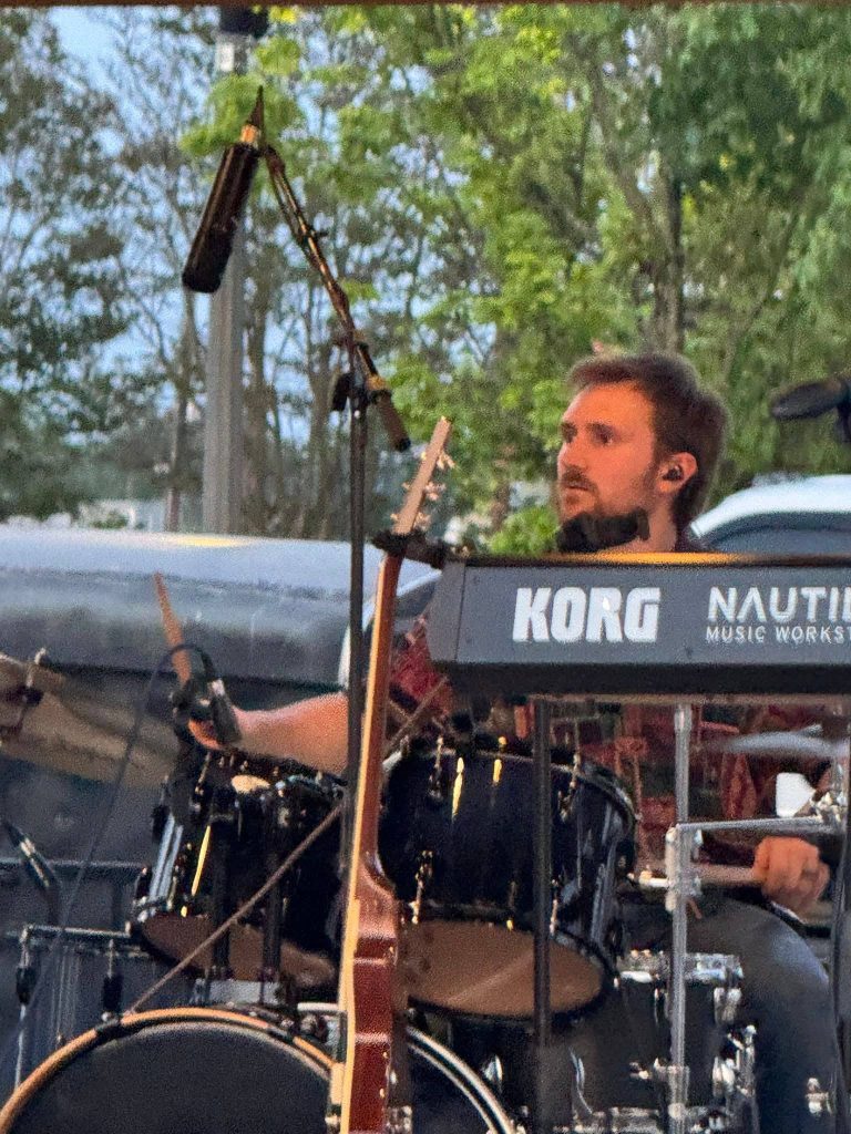 A musician playing drums and keyboards on an outdoor stage, with a microphone and various instruments visible, surrounded by trees in the background.