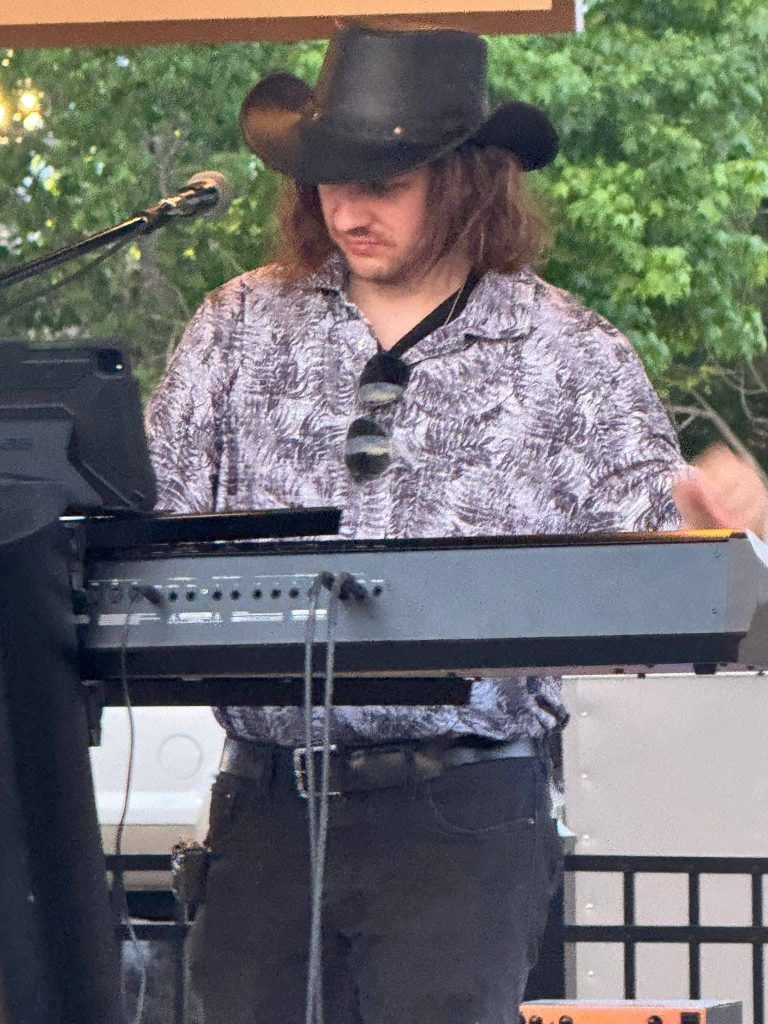 A man wearing a black cowboy hat and sunglasses plays a keyboard on an outdoor stage, with trees visible in the background.