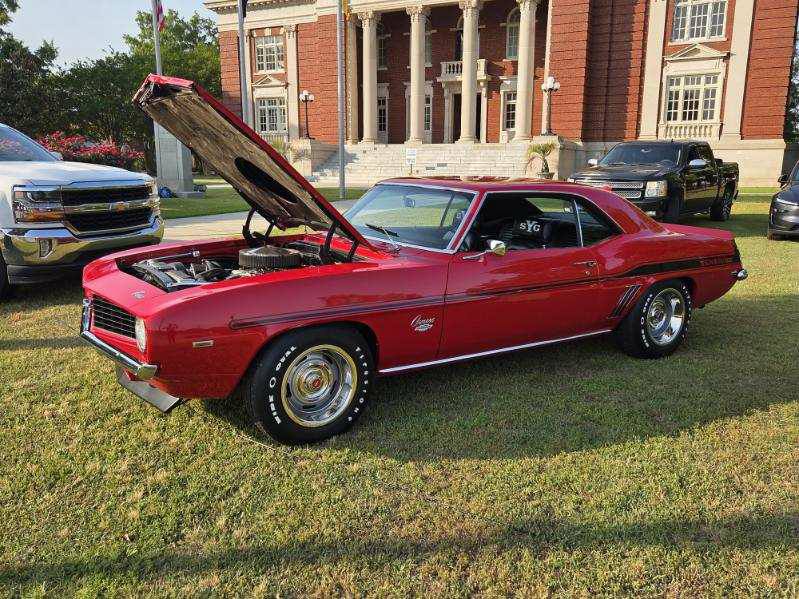 Classic red muscle car with hood open, parked on grass in front of a historic brick building, surrounded by modern vehicles.