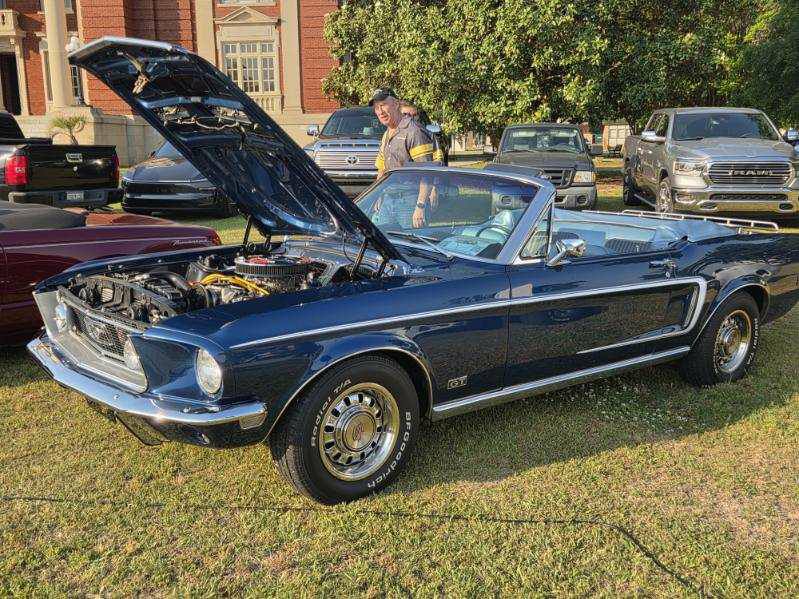 A classic blue convertible Ford Mustang with the hood open is displayed outdoors, with several cars and a building in the background.