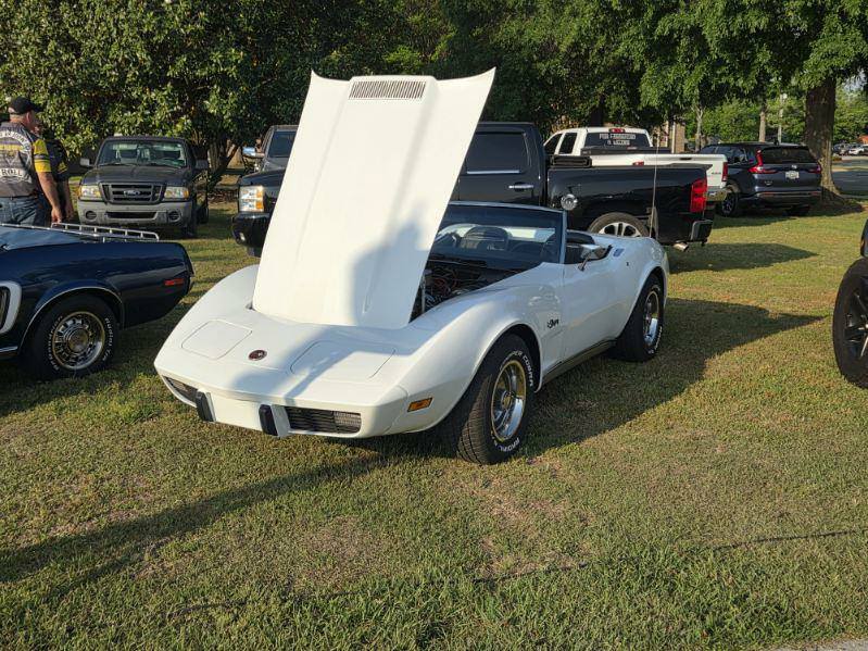A white classic convertible car with its hood open, parked on grass at a car show, surrounded by other vehicles and trees.