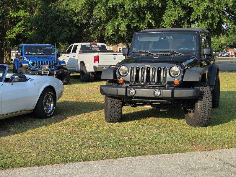 Several vehicles, including a black Jeep, blue Jeep, white pickup truck, and part of a white convertible, are parked on grass near trees on a sunny day.