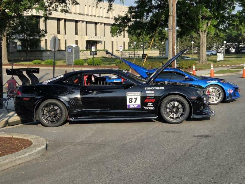 Two modified race cars parked outdoors with hoods open, showcasing sponsor decals and performance parts, surrounded by orange cones and trees near a large concrete building.