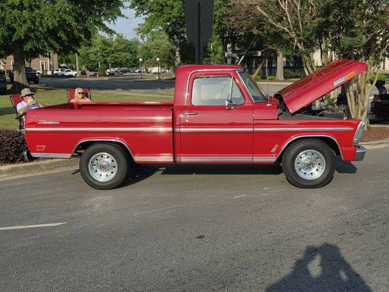 Red classic pickup truck with its hood open, parked on a street. Two people sit in lawn chairs nearby under trees on a sunny day.