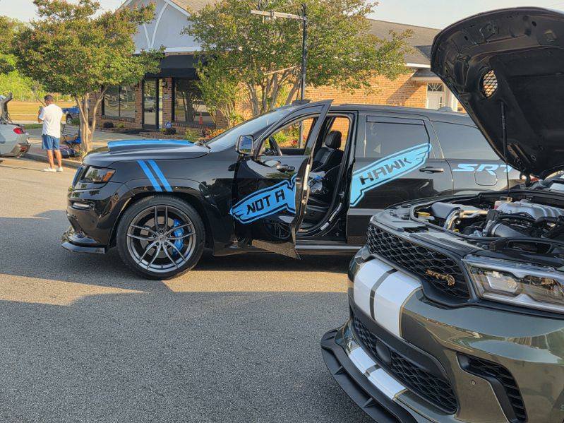 Black SUV with blue decals reading "Not a Hawk," driver door open, parked beside another car with hood open, trees and buildings in the background.