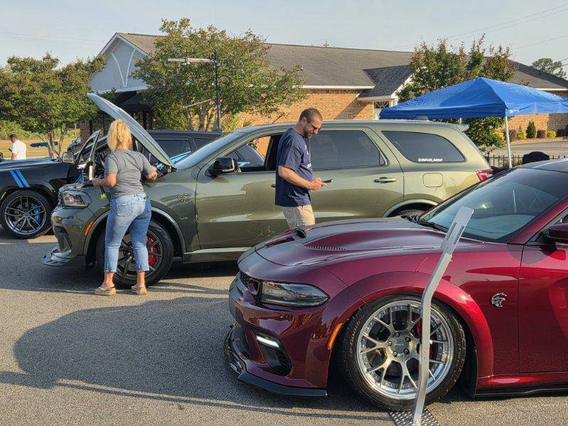 People examining cars at an outdoor car show, including a green SUV and a maroon sports car, with a tent and building in the background.