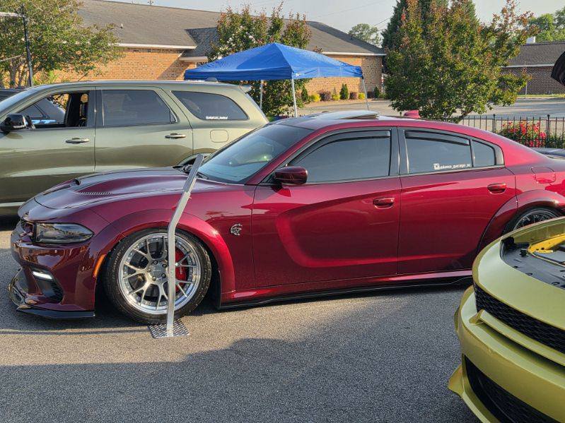 A red Dodge Charger with custom wheels parked at a car show, next to a green SUV and a yellow car, with a blue canopy in the background.