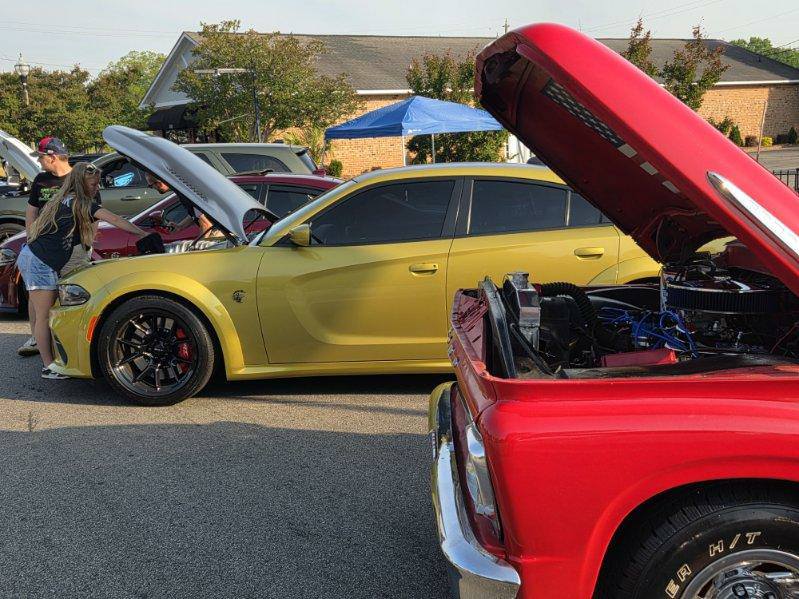 People view cars with open hoods at a car show, including a yellow sedan and a red truck, on a sunny day.