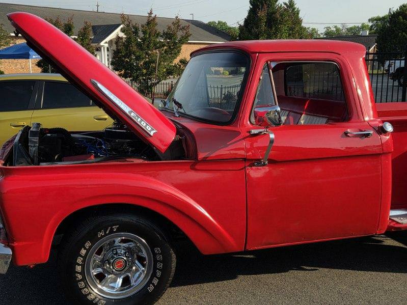 A classic red pickup truck with its hood open, parked on the street. The truck has shiny chrome wheels and a vintage design.