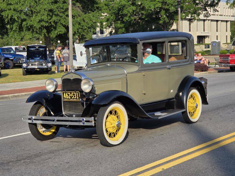 A vintage olive green car with yellow wheels drives down a street during a classic car show, with people and other cars visible in the background.