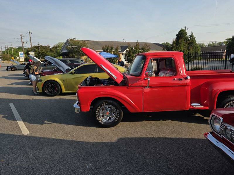 Classic cars and trucks with open hoods are lined up at a car show in a parking lot, with people viewing them.