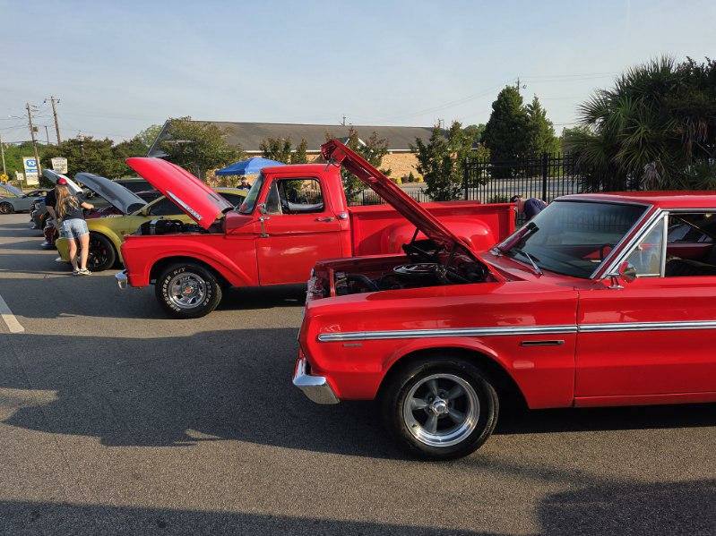 Classic cars and a red vintage truck with open hoods are parked in a line at an outdoor car show.