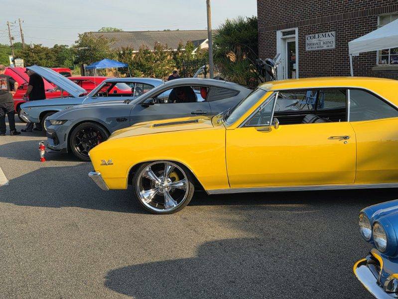 Several classic and modern cars are parked in a row at an outdoor car show, with people walking and inspecting vehicles.