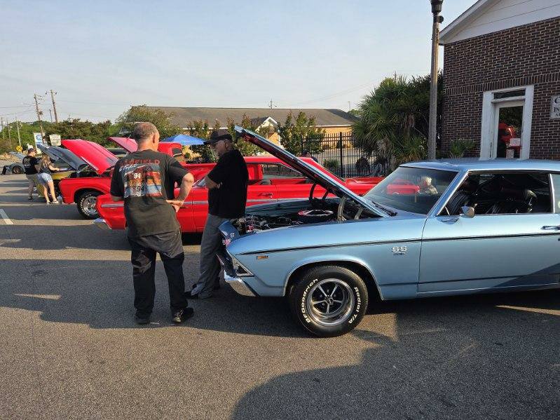 People talk near classic cars with open hoods at an outdoor car show, featuring a light blue Chevrolet SS and a red vehicle in the background.