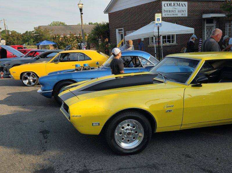 Classic cars, including a yellow Chevrolet Camaro, parked at a car show with people admiring the vehicles outside a brick building.