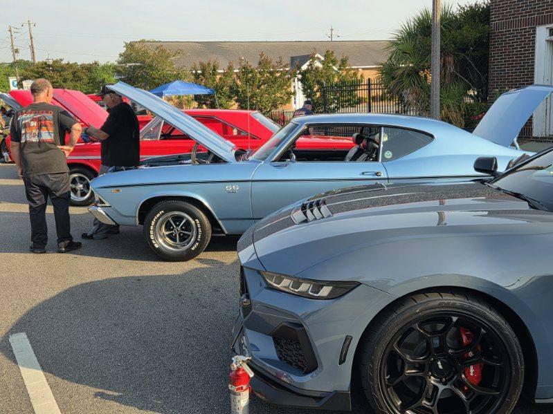 Several classic and modern cars with open hoods are parked at an outdoor car show, with people standing and talking beside them.
