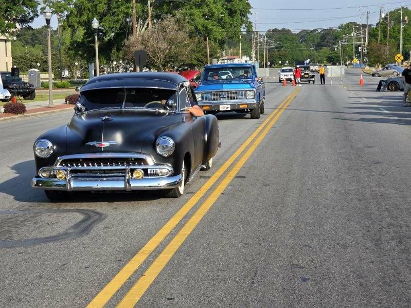 Classic black car and blue pickup truck drive down a city street lined with trees, while people and traffic cones are visible in the background.