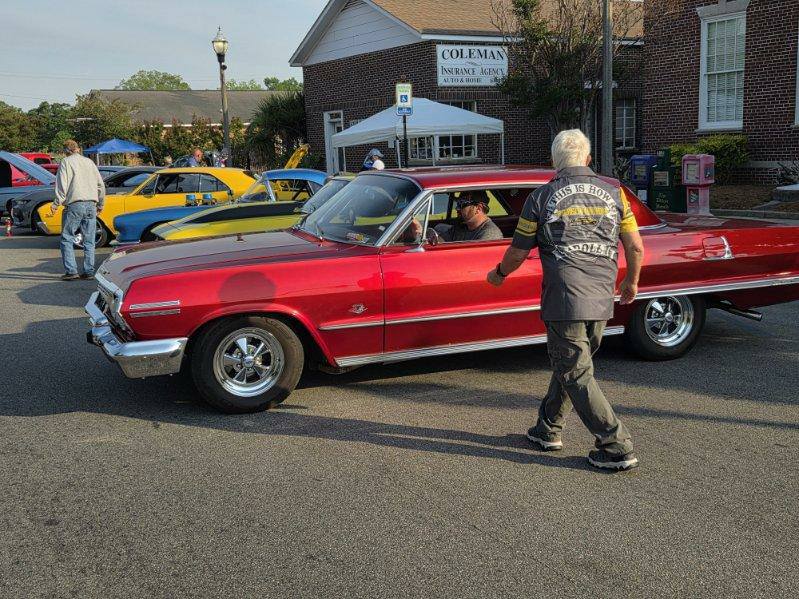 A classic red car at a car show with people around, set in front of a brick building labeled "Coleman Insurance Agency."