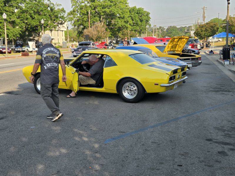 A man sits in a yellow classic car with black stripes at a car show, while another man walks nearby. Other classic cars are parked in the background.