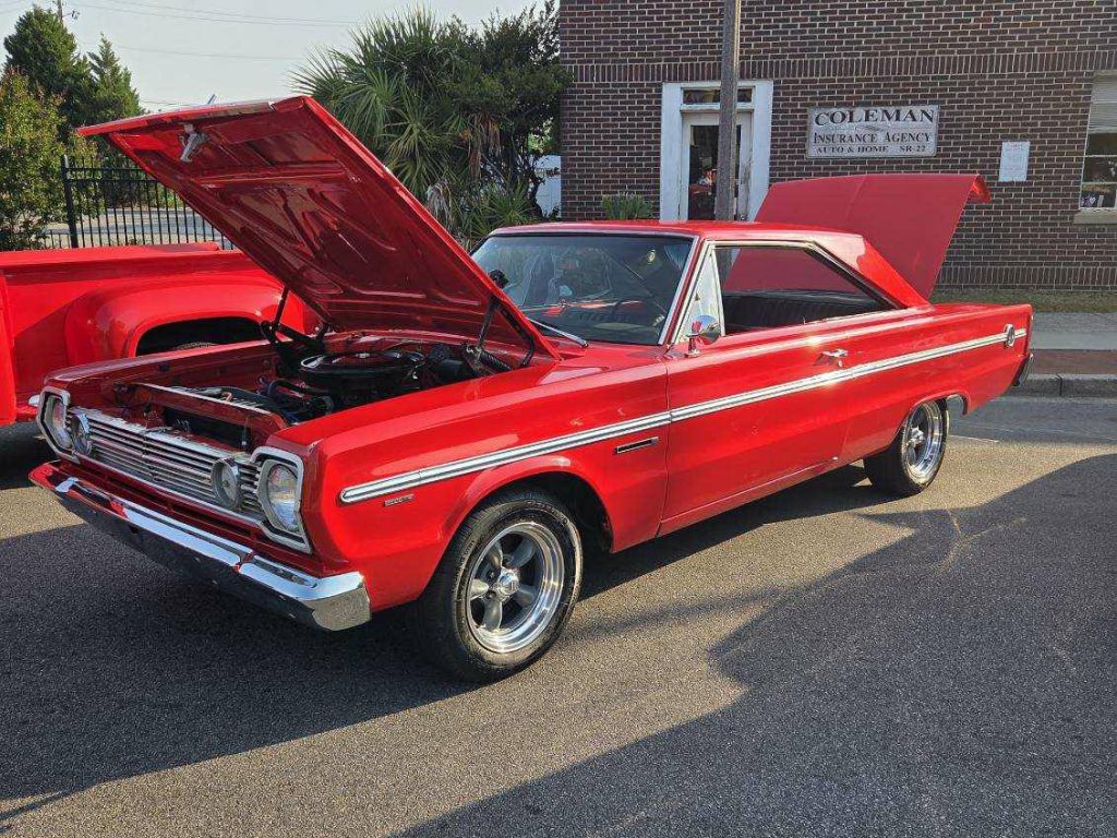 A classic red muscle car with its hood open, parked on a street in front of a brick building and insurance agency sign.